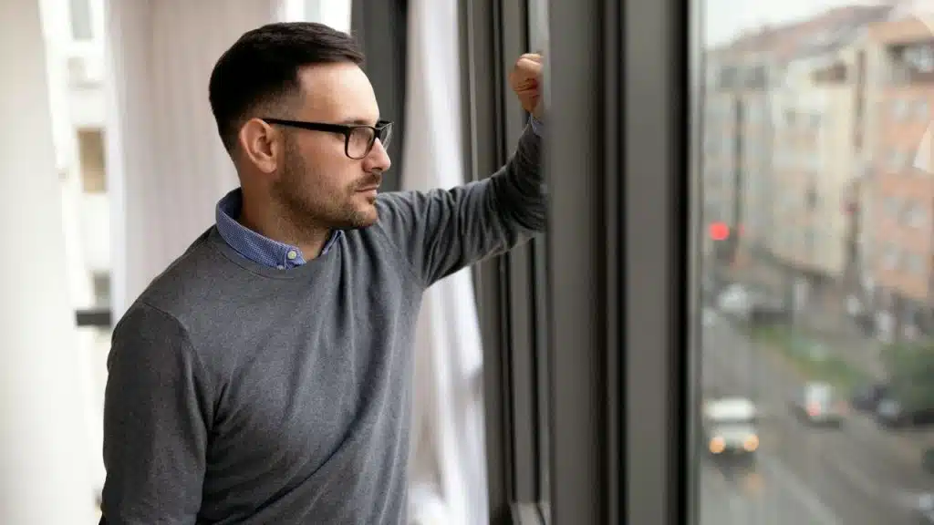 A man with glasses and a beard, wearing a grey sweater, looks out a window.