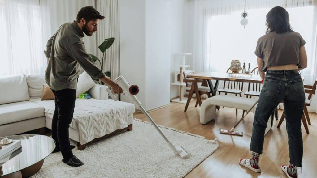 A man vacuums a rug in a living room while a woman mops the floor.