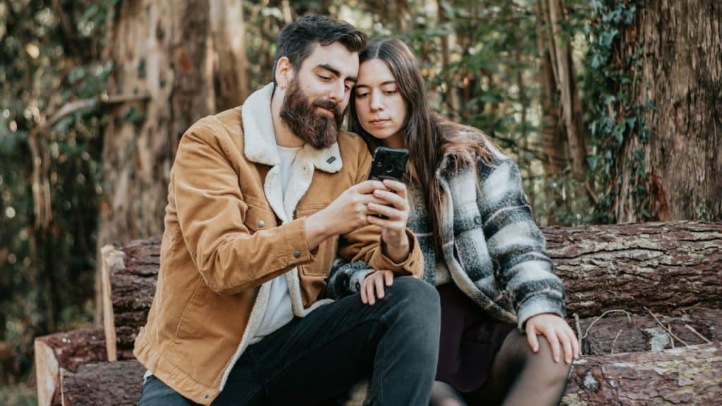 A focused couple sitting on a log, looking intently at a smartphone.