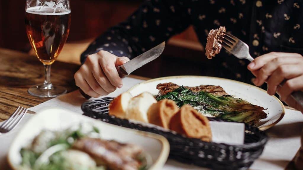 A person eating steak with greens, bread, and a glass of beer.