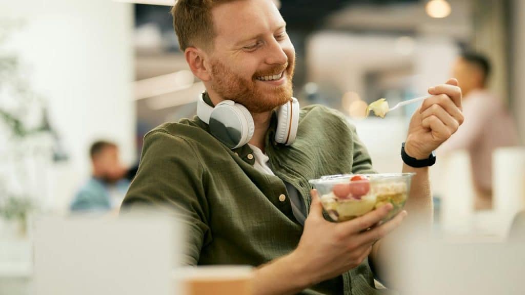 A smiling man with headphones eating a salad in a modern indoor setting.