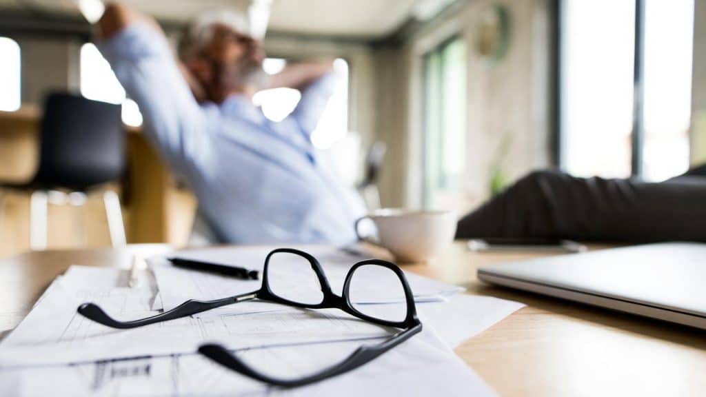 A person's glasses resting on papers with a relaxed man in the background.
