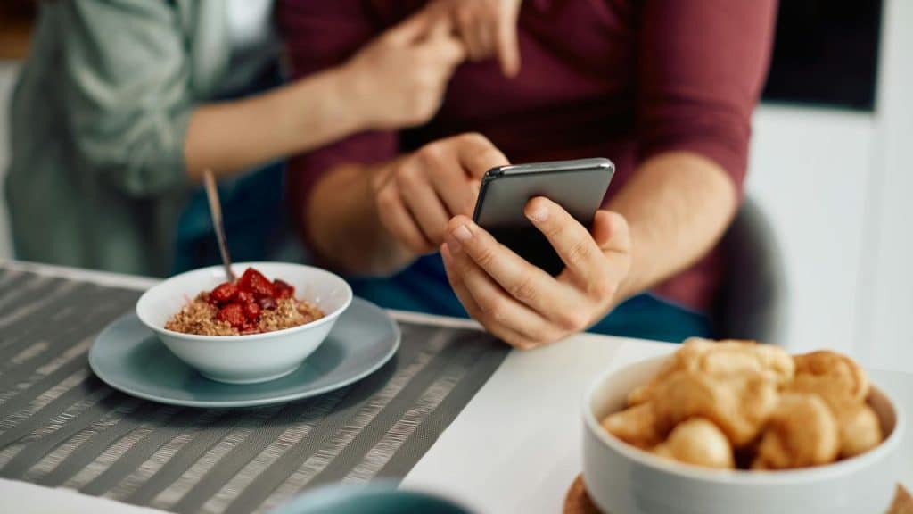 A person's hand holding a phone at the breakfast table with cereal and pastries.