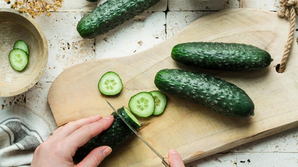 A person's hands slicing cucumber on a wooden cutting board.