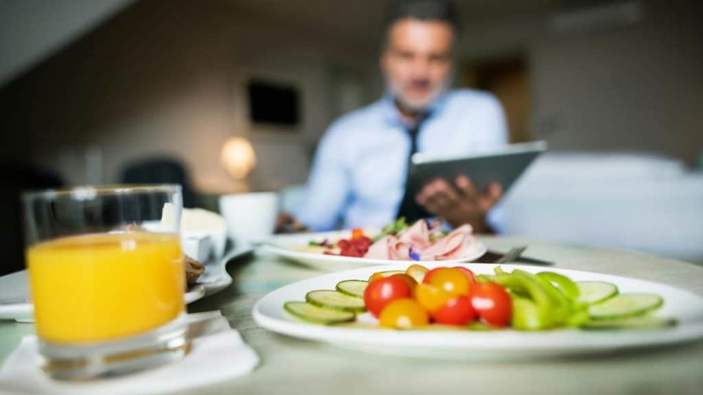 A man eating breakfast while using a tablet, with juice and vegetables in the foreground.