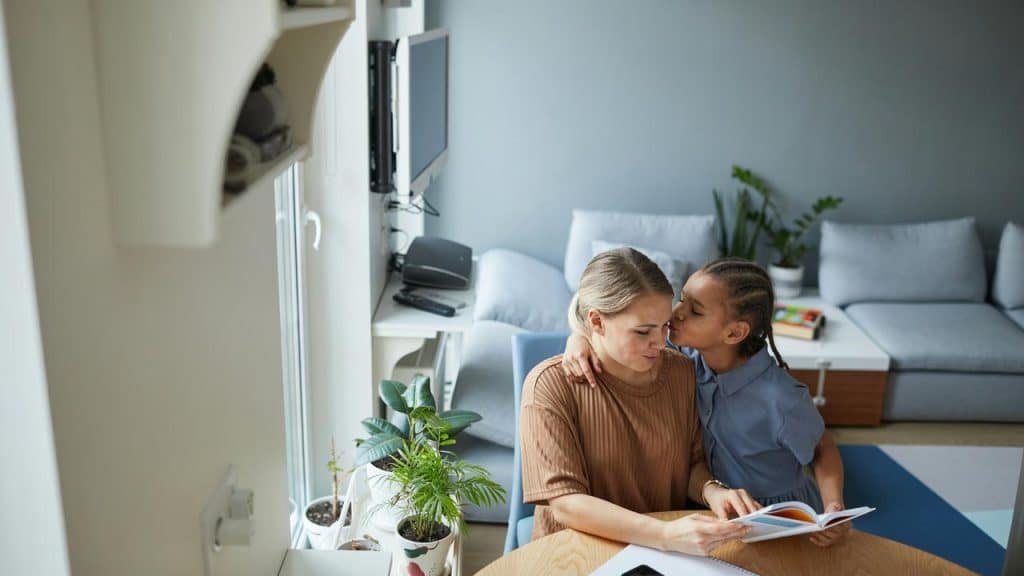 A child kissing their mother while she reads at a table.