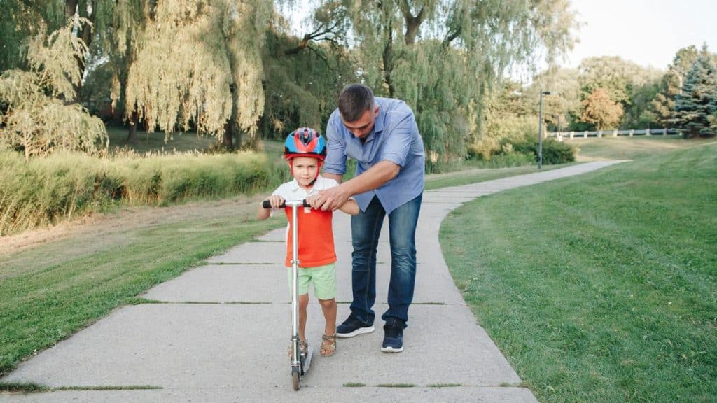 A father helps his son ride a scooter.