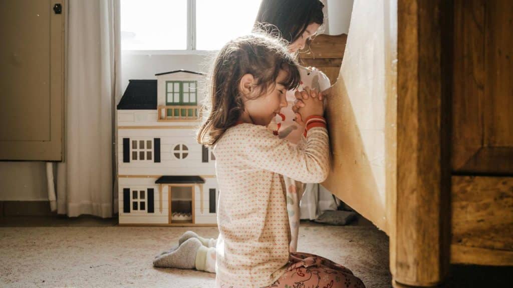 A child kneels in prayer beside a bed.