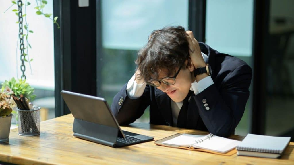 A man in a suit sits at a desk with his hands in his hair, looking stressed.