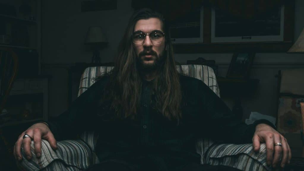 A man with long hair, a beard, and glasses sits in a striped armchair.