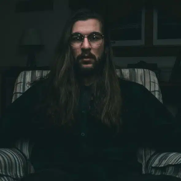 A man with long hair, a beard, and glasses sits in a striped armchair.