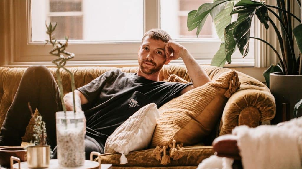 A man with short hair and a beard reclines on a yellow couch, looking at the camera.