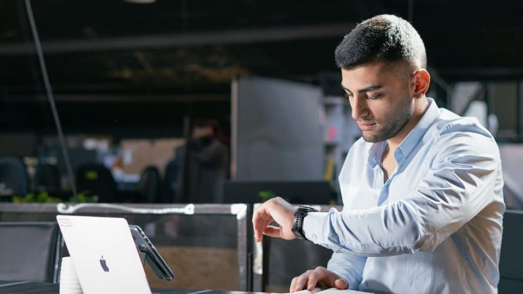 A man in a blue shirt looks down at the watch on his wrist while at a desk.