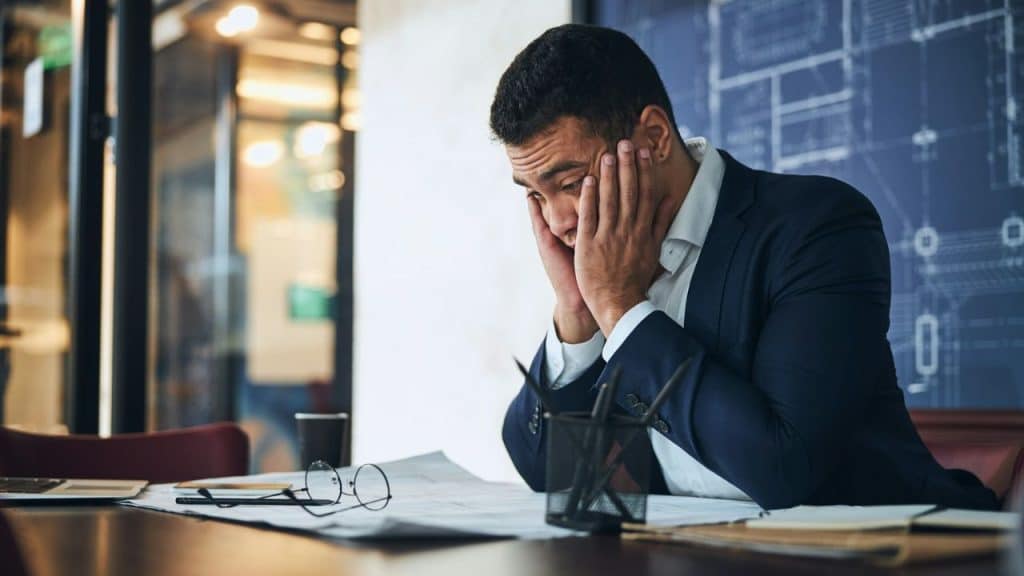 A man in a suit sits at a desk with his head in his hands, looking stressed.