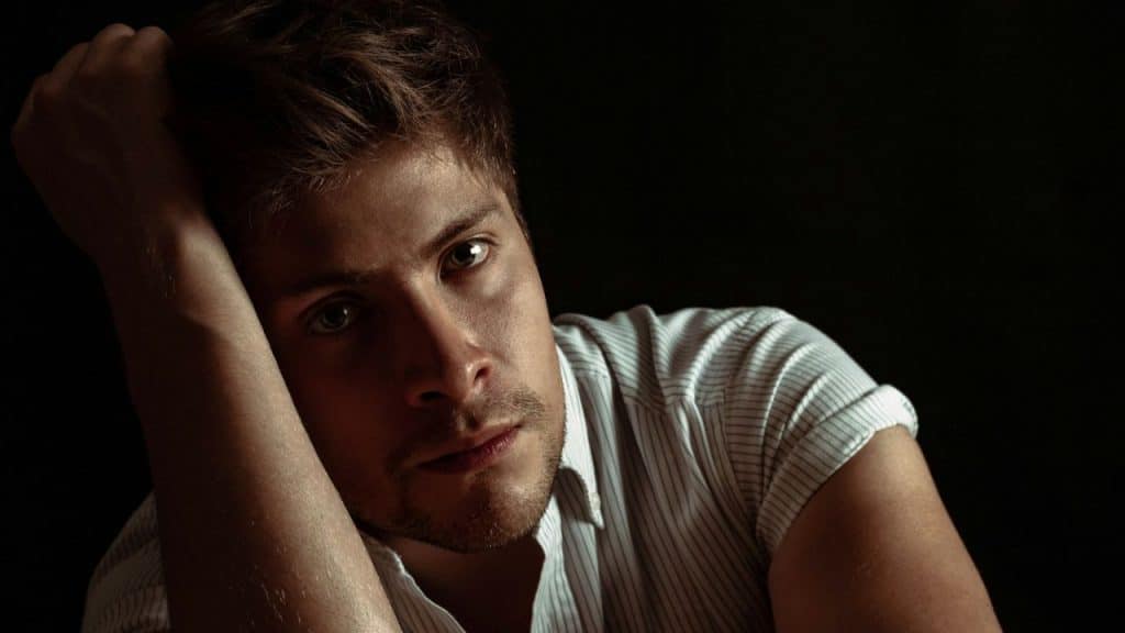 A man with light hair and a beard rests his head on his arm, looking at the camera.
