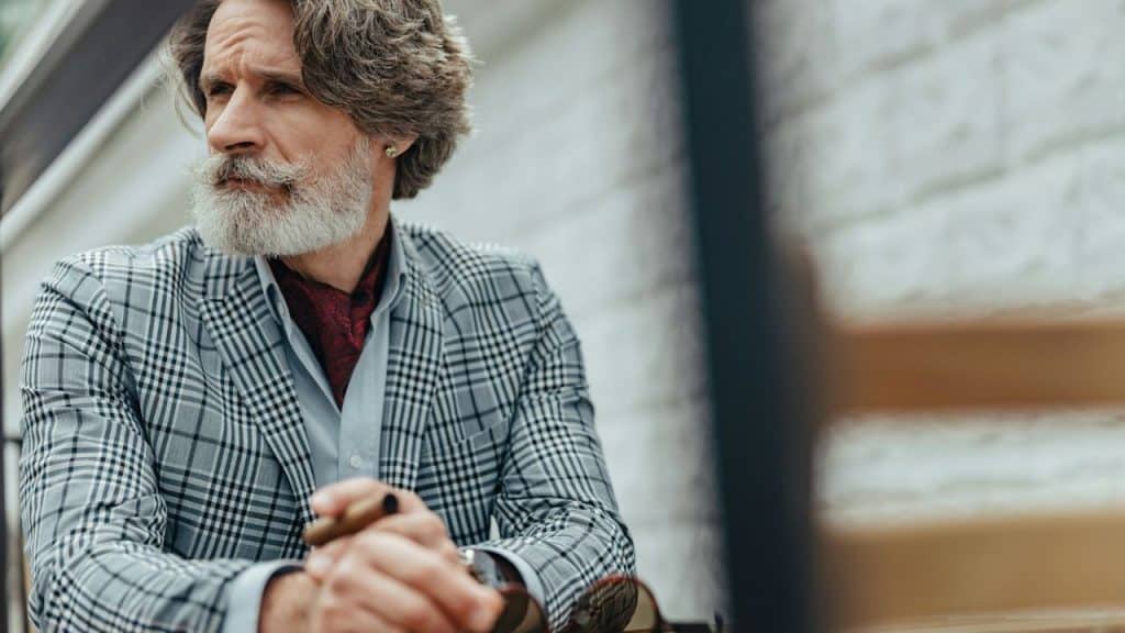 A well-dressed older man with gray hair and a beard sits outdoors, holding a cigar.