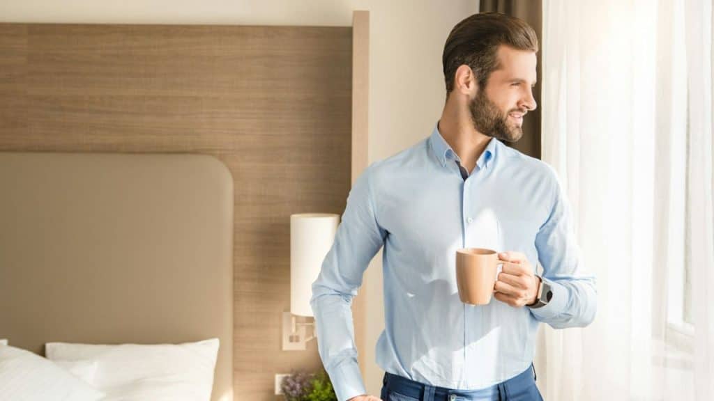 A smiling man in a blue shirt holds a mug and looks out a window.