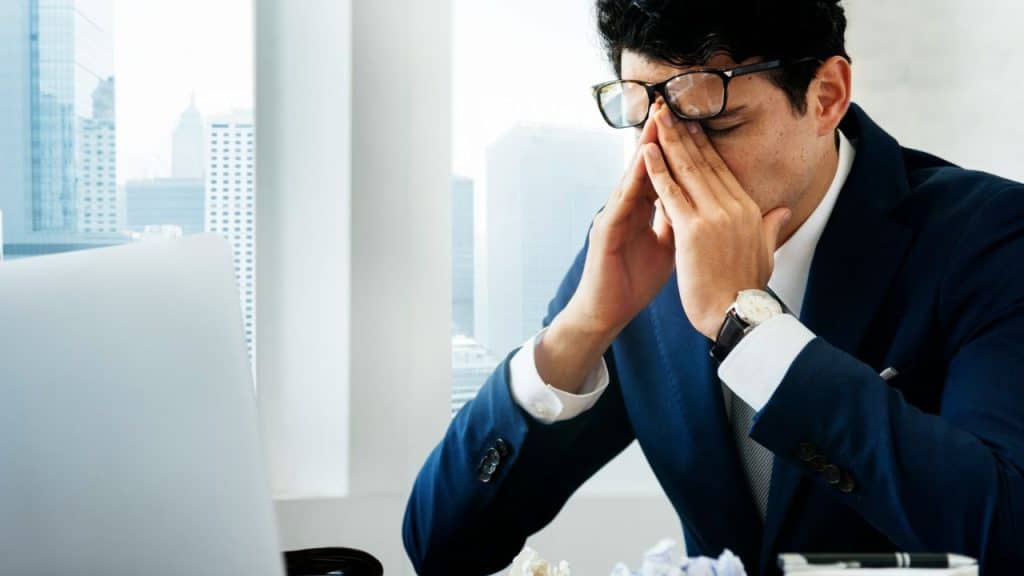 A man in a suit and glasses sits at a desk, rubbing his eyes in frustration.
