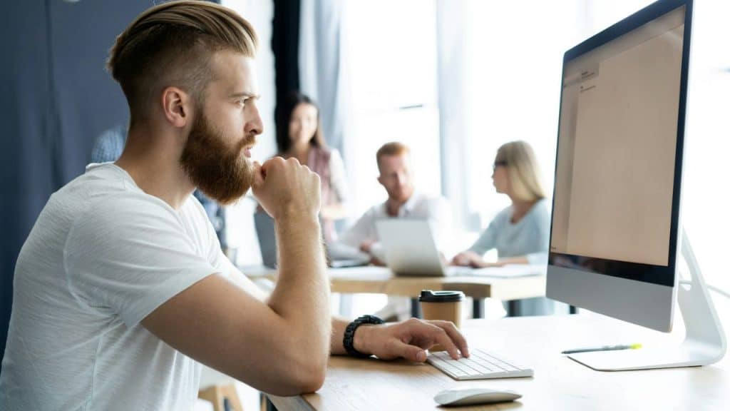 A man with a beard sits at a desk, looking at a computer screen in an office.