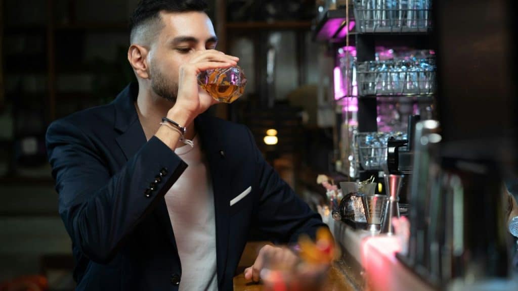 A man in a navy jacket sips a drink at a bar counter.