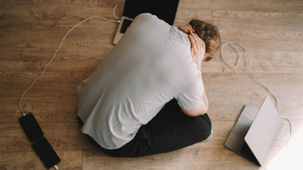 A person sits on a wooden floor, huddled and holding their head, with a laptop and phone nearby.