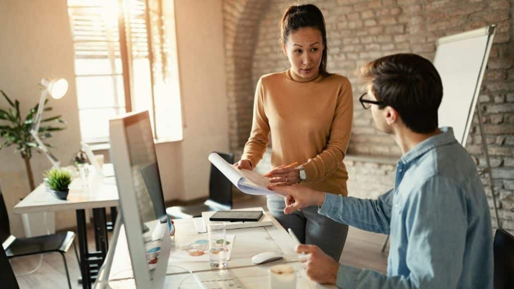 A woman stands over a man at a desk, pointing at papers on a computer monitor.