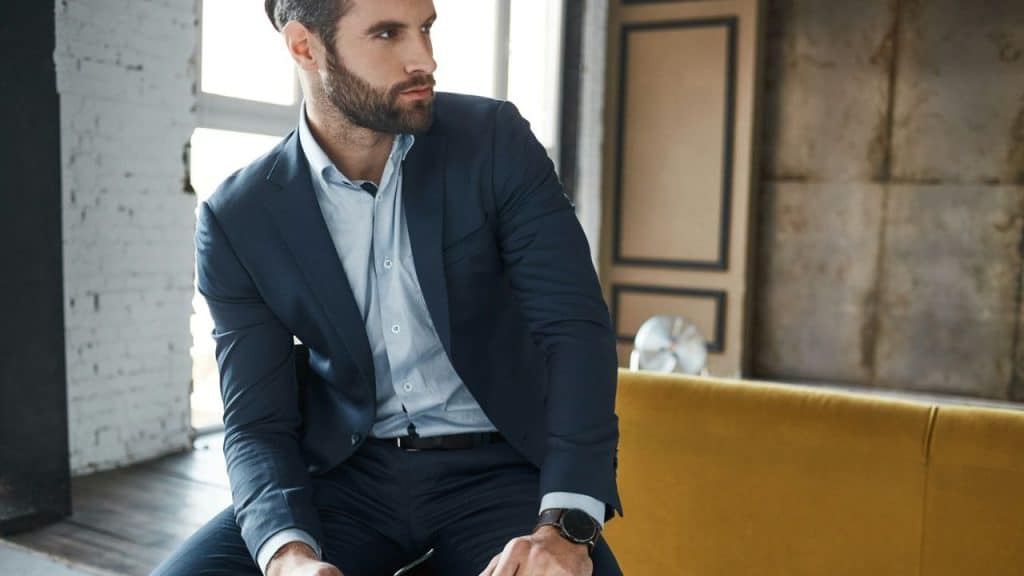 A man with a beard in a navy suit sits indoors, looking to his left.