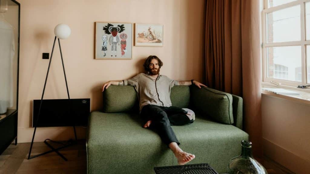 A man with a beard and long hair sits barefoot on a green couch, looking at the camera.