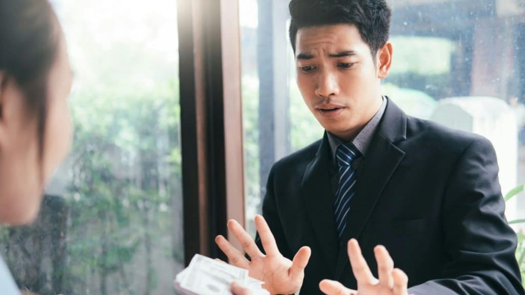 A man in a suit looks stressed as he gestures with his hands, facing a person holding money.
