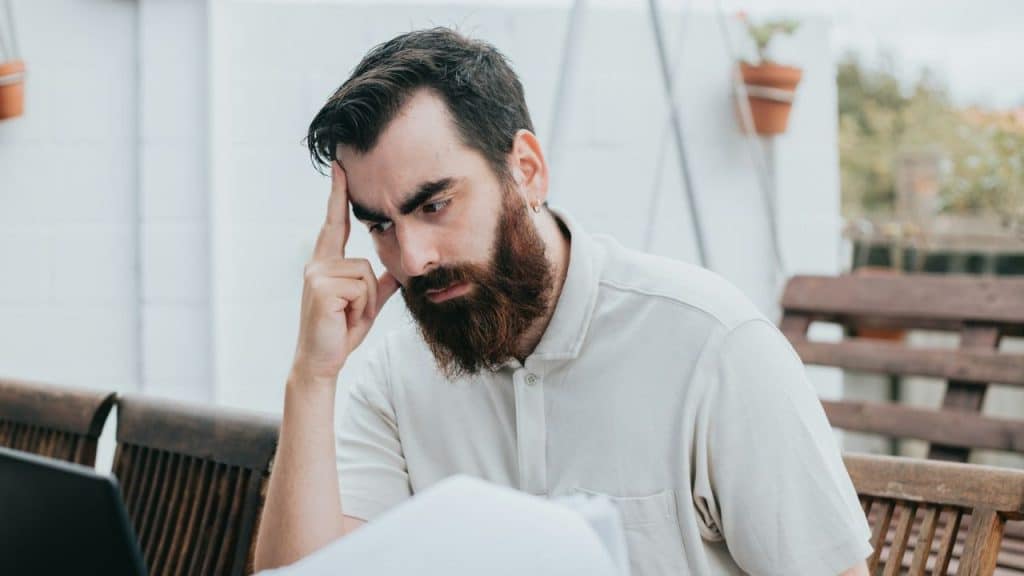 A man with a full beard and a white polo shirt sits with his hand on his head, looking stressed.