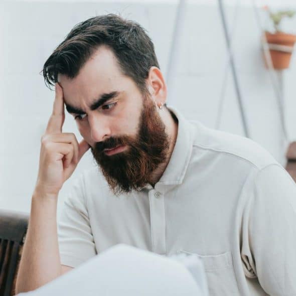 A man with a full beard and a white polo shirt sits with his hand on his head, looking stressed.