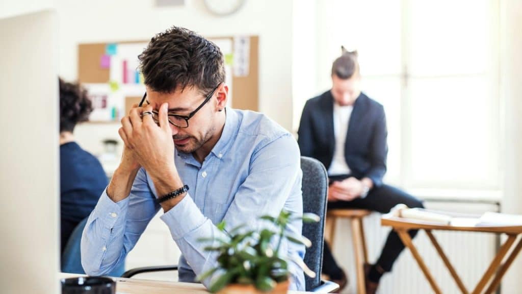 A man with glasses at a desk holds his head in his hands, looking stressed.
