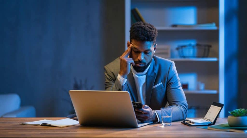 A man sits at a desk with his head in his hand, looking at his phone.