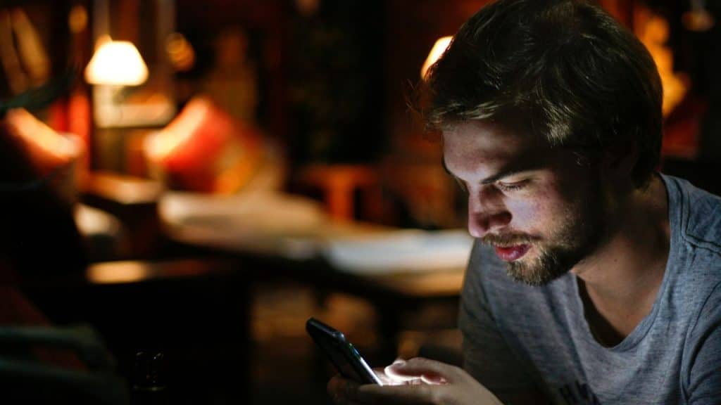 A man with a beard looks down at his phone in a dimly lit room.