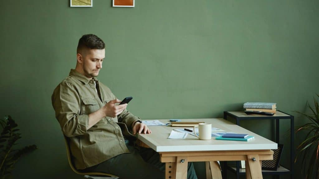 A man with short hair sits at a desk, looking at his phone.