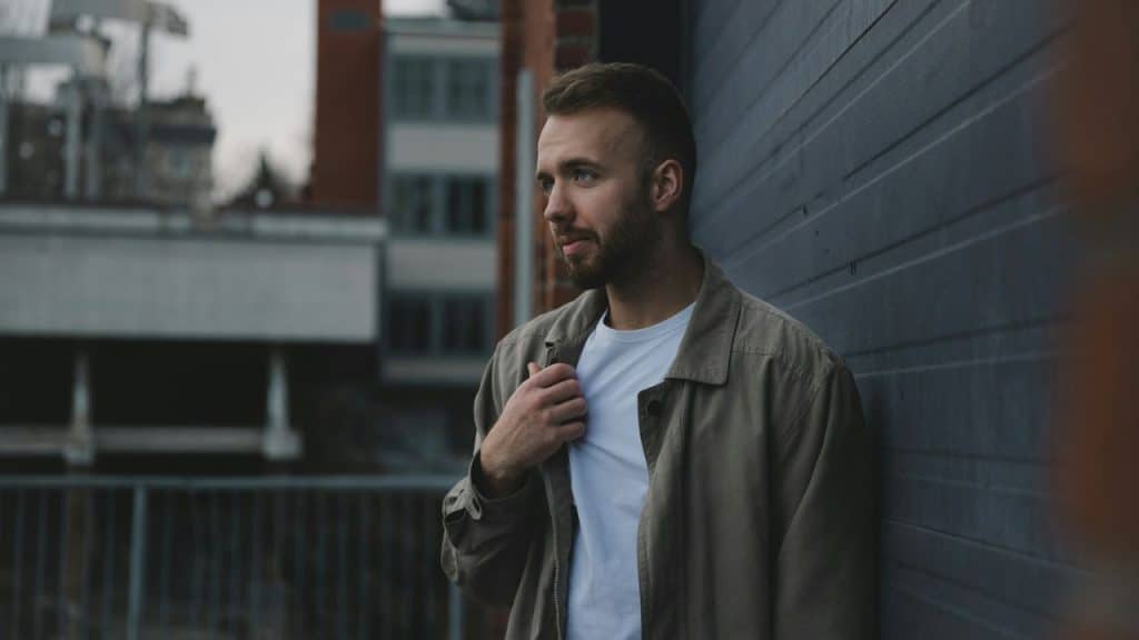 A man with a beard leans against a wall, adjusting his jacket while looking to the side.