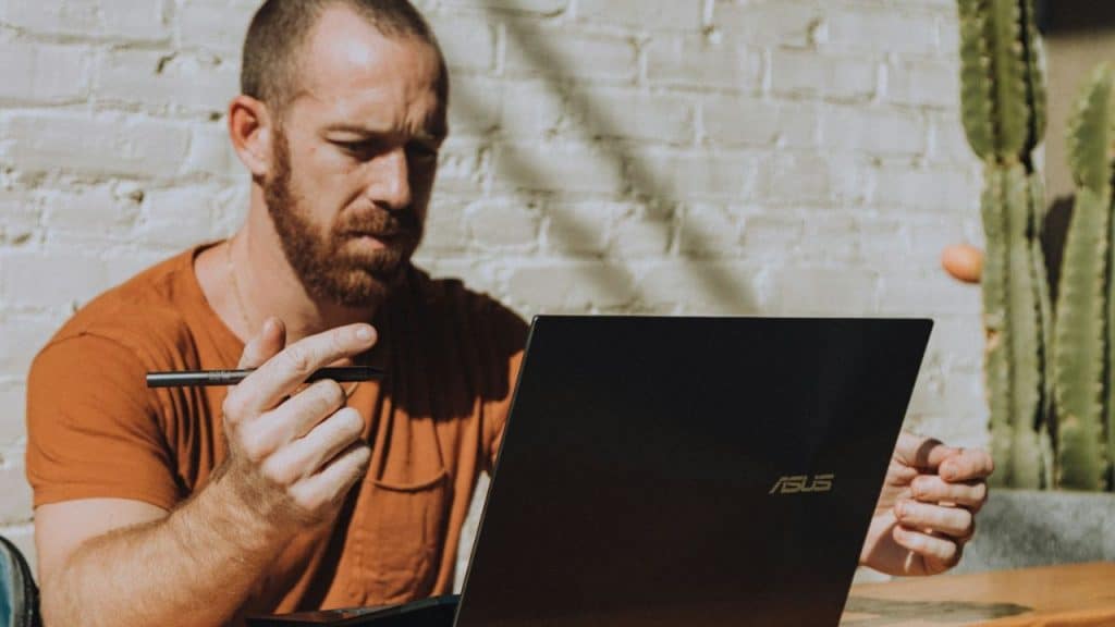 A man with a beard holds a stylus, looking intently at his laptop screen.
