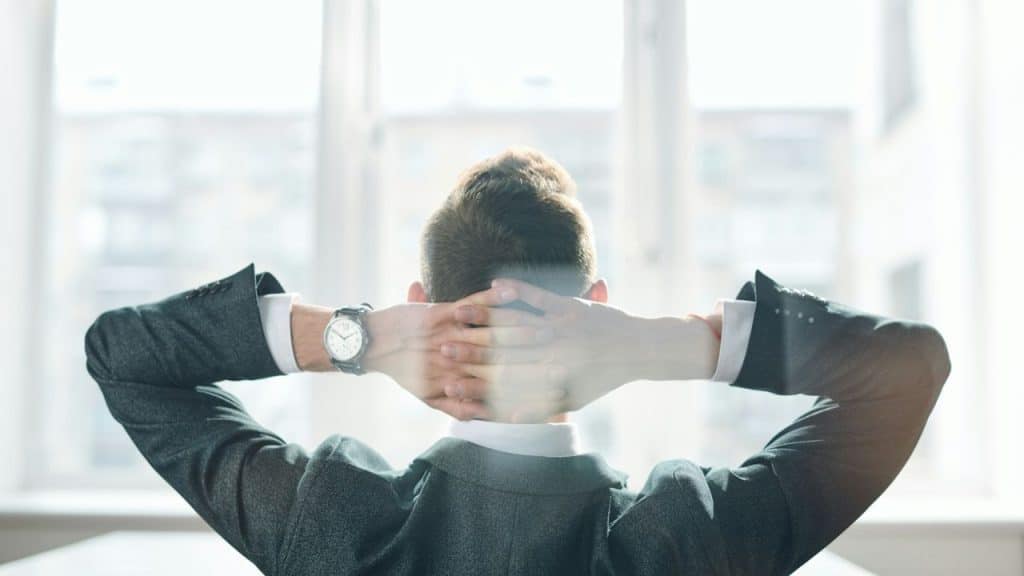 A man in a gray suit sits with his hands behind his head, looking out a window.
