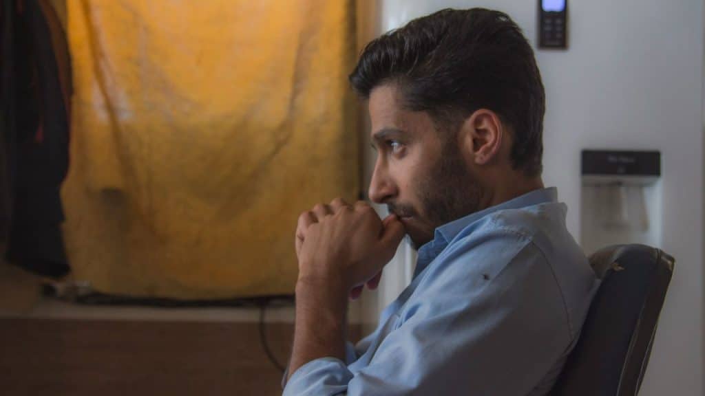 A man in a light blue shirt sits with his hands clasped together, looking concerned.
