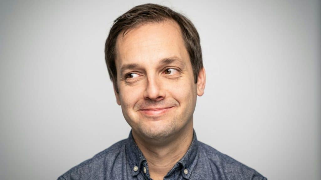 A man in a blue shirt with a smirk on his face looks to the side.