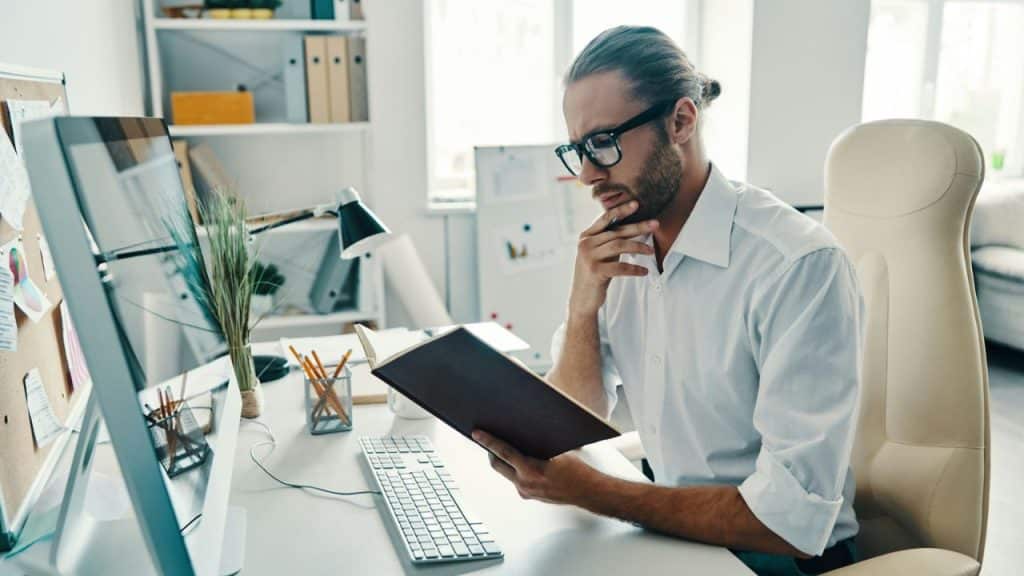 A focused man with glasses reads a book at a modern desk.
