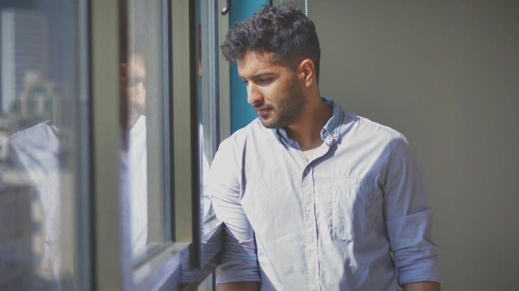 A thoughtful man looks down while standing near a window.