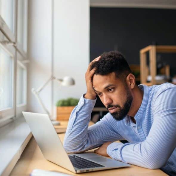 A worried man sits at his desk, staring at his laptop screen.