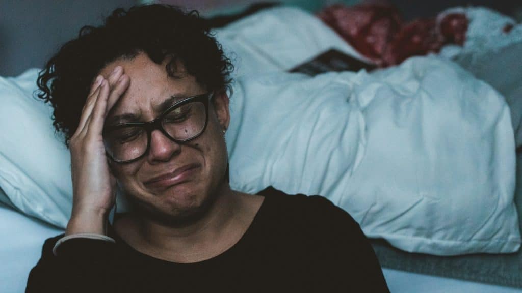 A crying woman with glasses clutches their head in a dimly lit room.