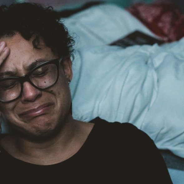 A crying woman with glasses clutches their head in a dimly lit room.