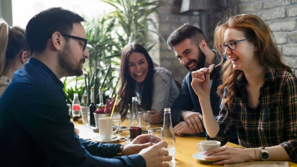A man in glasses looks to the side while others at the table laugh.