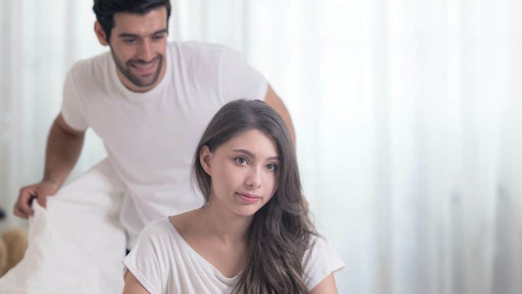 A smirking man holds a blanket behind a woman who looks ahead.