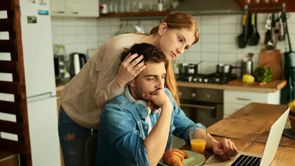 A concerned woman stands behind a man who is looking at a laptop at a kitchen table.