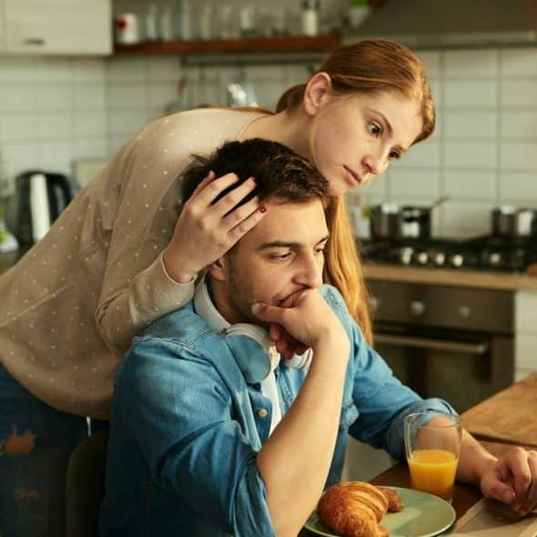 A concerned woman stands behind a man who is looking at a laptop at a kitchen table.