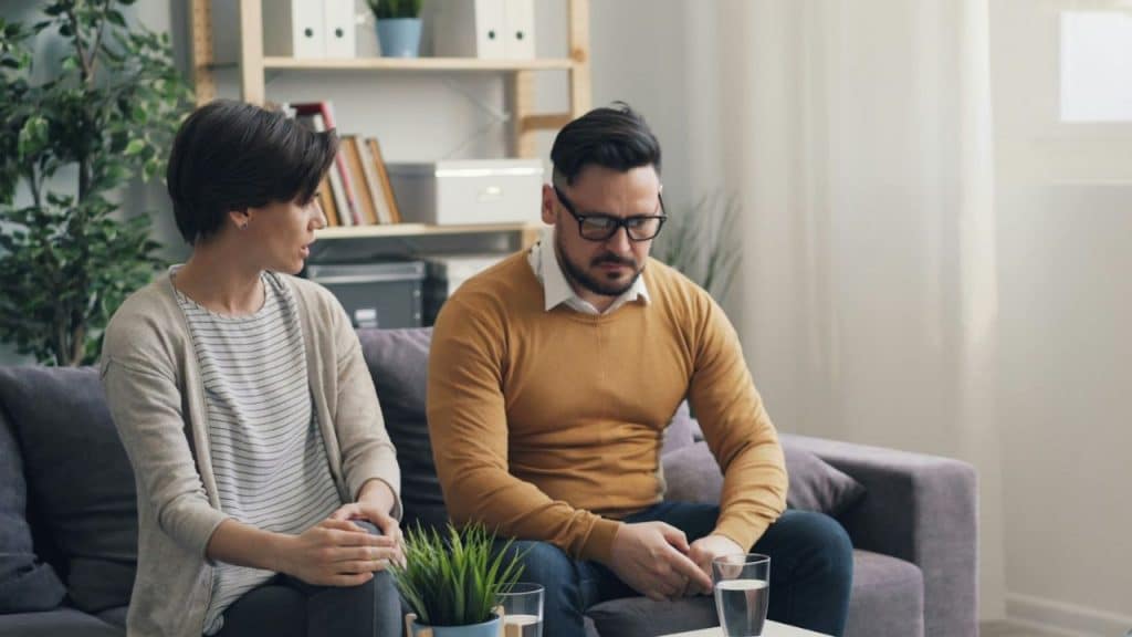 A concerned woman looks at a sad man sitting with his hands clasped.